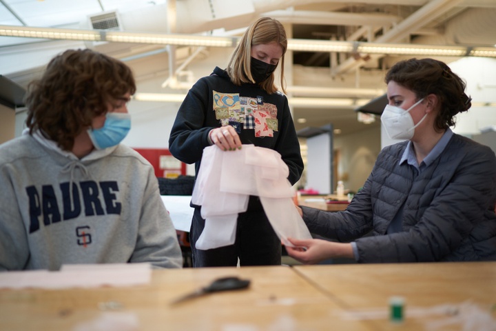 Two students and a faculty member gather around a studio desk, holding and looking at a white fabric garment.