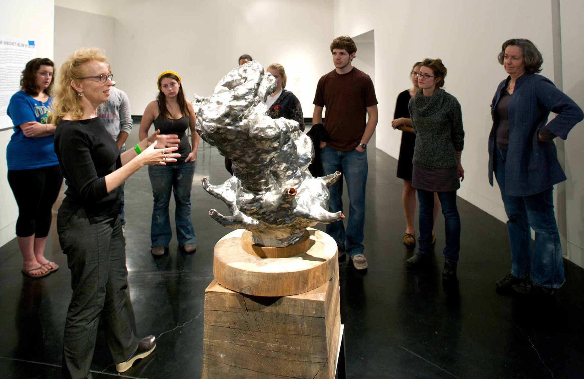 A group of people gather around an abstract, silver sculpture on a wooden pedestal.
