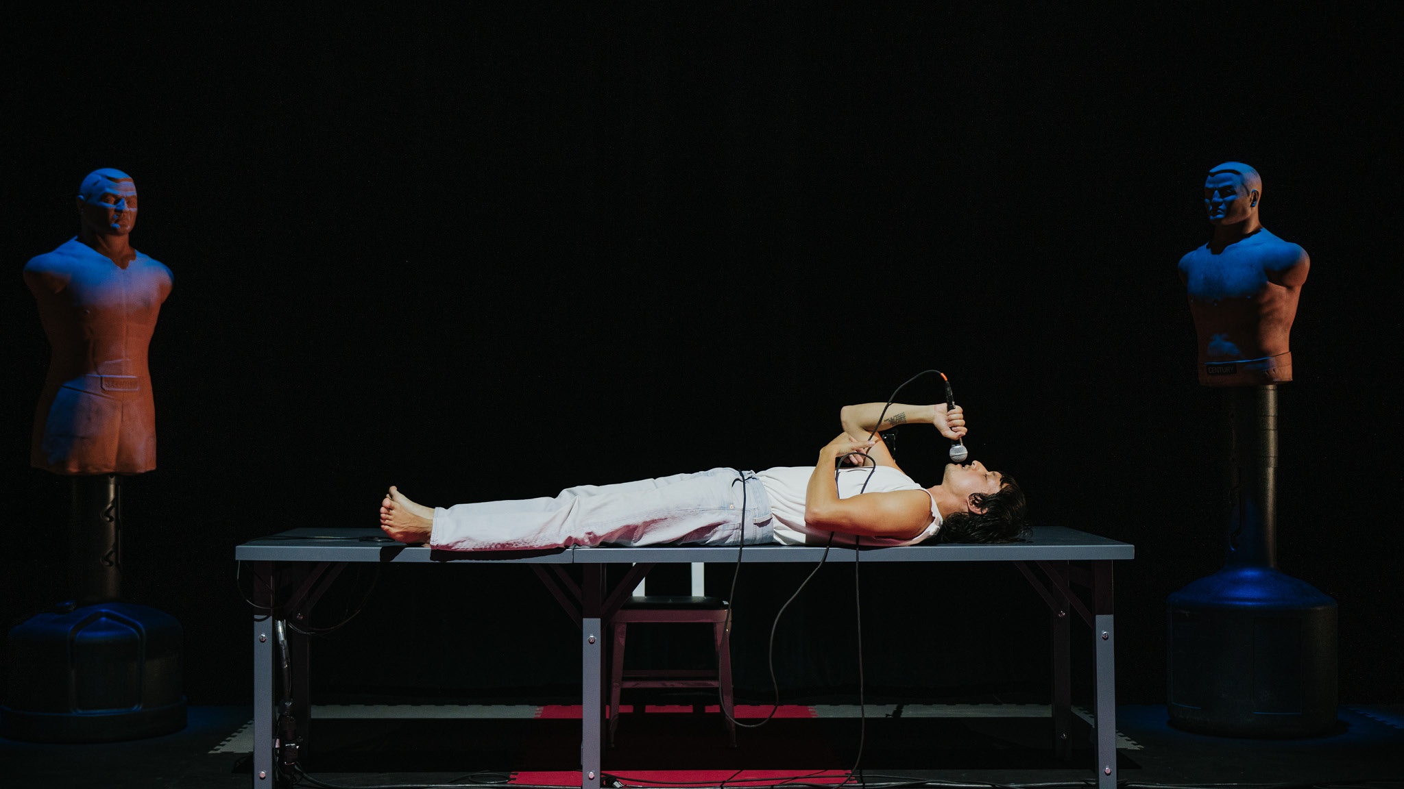 The artist Kyle Dacuyan lies on a folding table during a performance. The table is positioned on a dark stage between two sparring dummies. Kyle wears light-colored clothing and lies on his back holding a microphone up to his mouth. 