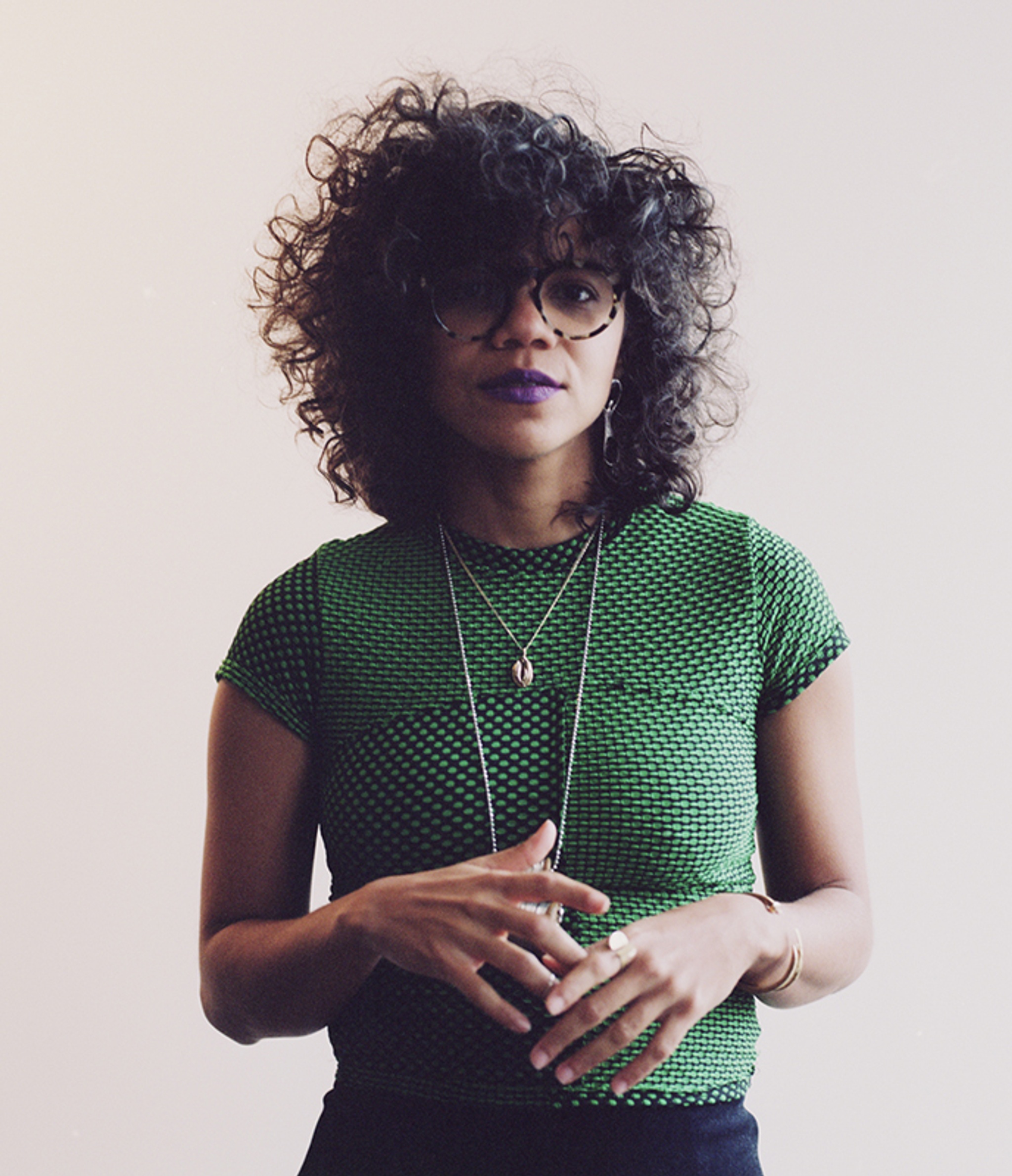 A portrait of Lizania Cruz, a Dominican artist who has curly hair that she wears over her forehead, slightly covering her eyes. She poses with her hands brought together in front of her waist and wears a green short sleeve shirt, long necklaces, glasses. She looks at us. Photo by Guarinex Rodriguez. 