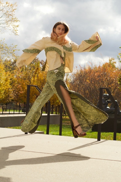 Person makes a running pose midair wearing wide-leg green brocade trousers with slits up to the hip and a cream-colored button-down top with wide sleeves and detailing in the same green brocade.