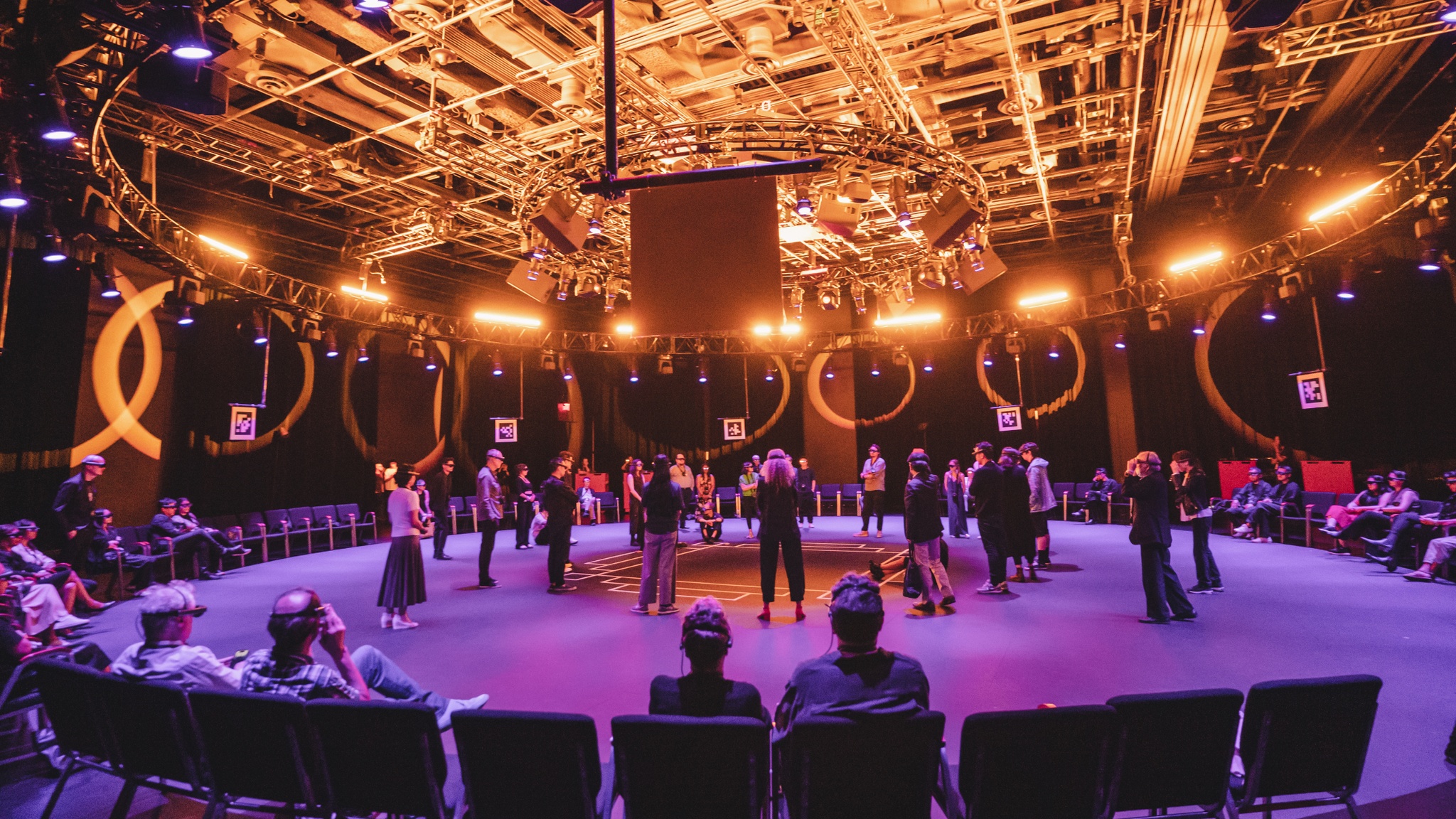In a theater space arranged with a wide circle of chairs facing inside the circles, audiences gather in the center of the space, standing and crouching. They were headsets that look like googles over their eyes. The space is lit in purple light that illuminates the floor and a bright golden light that reveals the infrastructure of pipes and ducts in the ceiling.