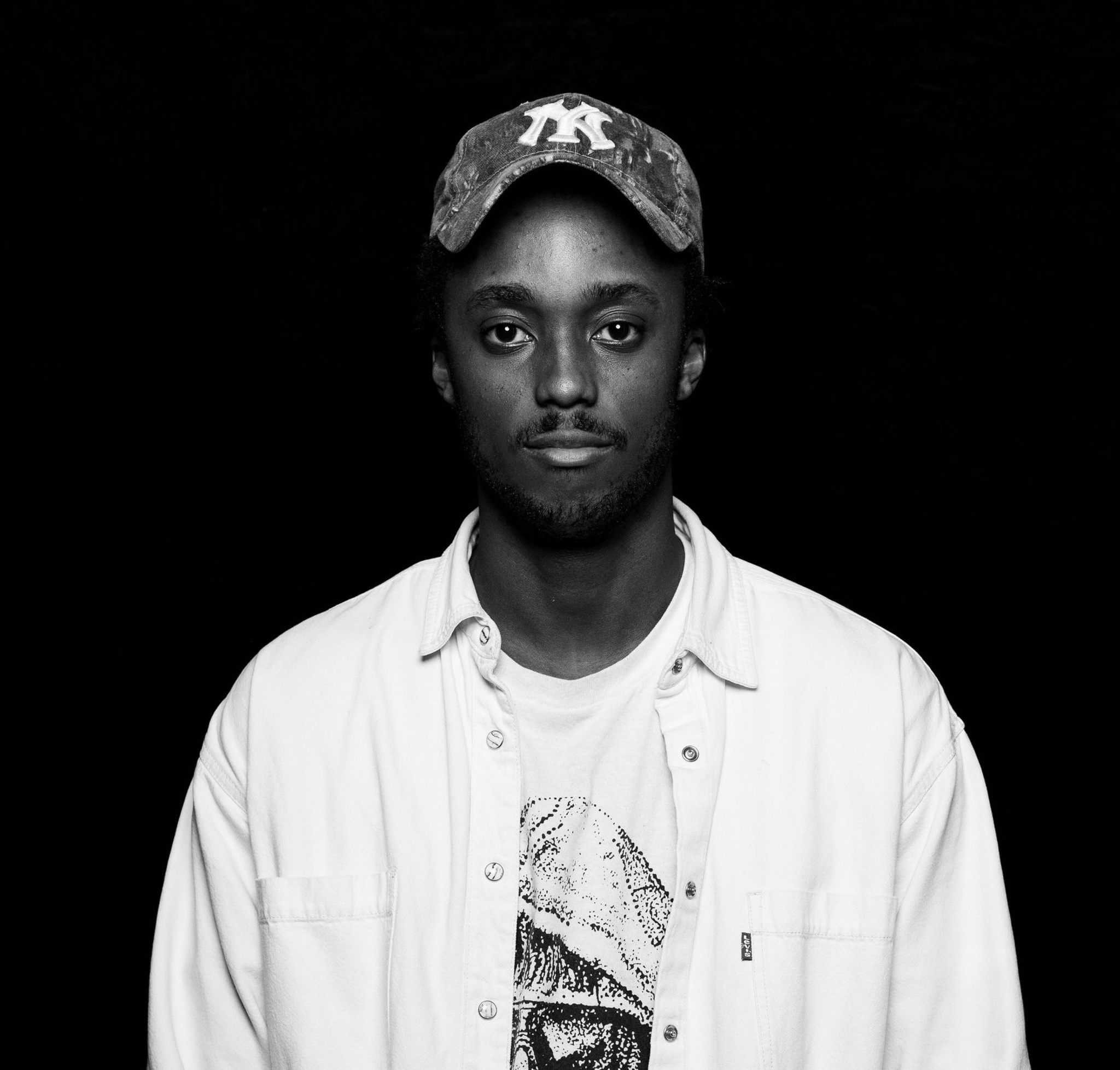 This is a black-and-white headshot of Nile Harris. They pose against a dark background. They are wearing a baseball cap and a white button down shirt open over a tshirt.