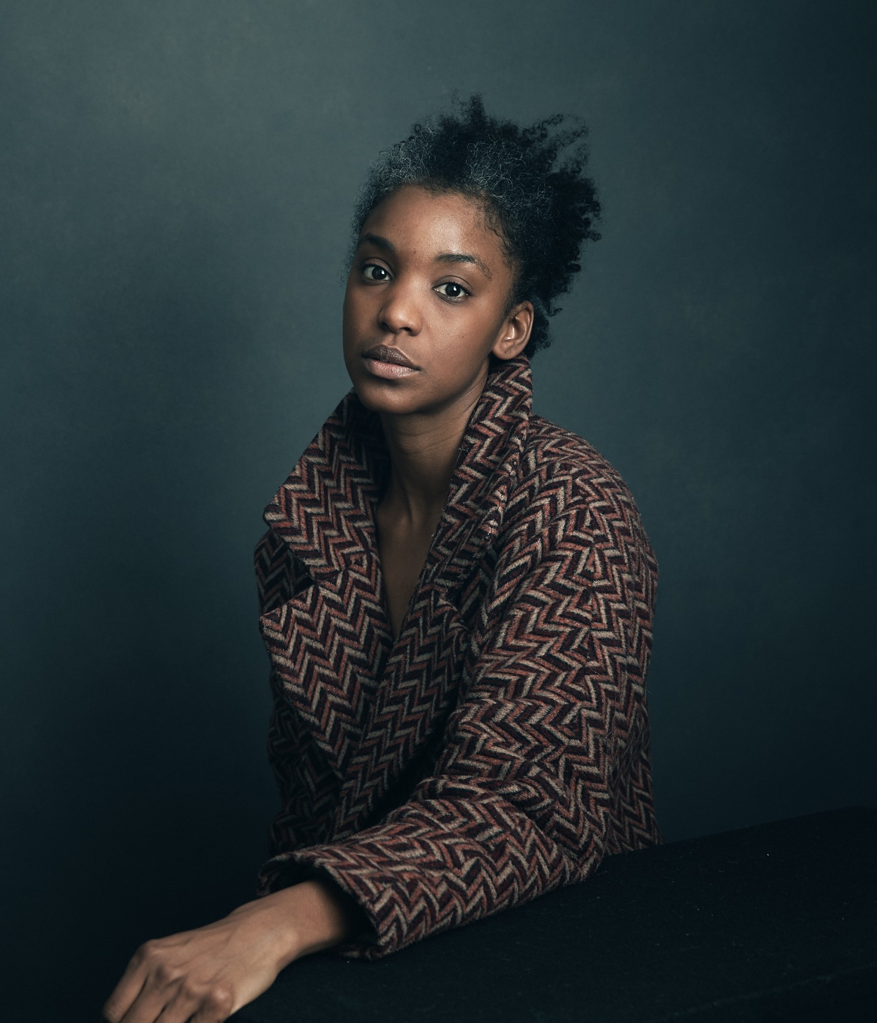 A portrait of the artist Ayesha Jordan sitting against a dark gray background with one arm outstretched and wearing chevron-patterned jacket