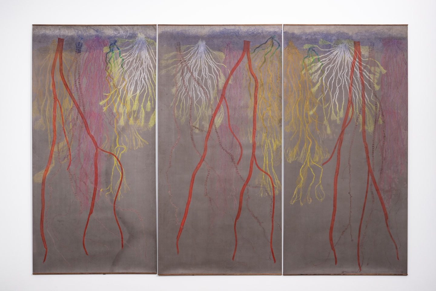 Large relief prints across three vertical panels depict prairie root systems in red, white, yellow, green, and purple. The printed lines vary from thick to delicate as they branch and descend over a muted gray-brown background.