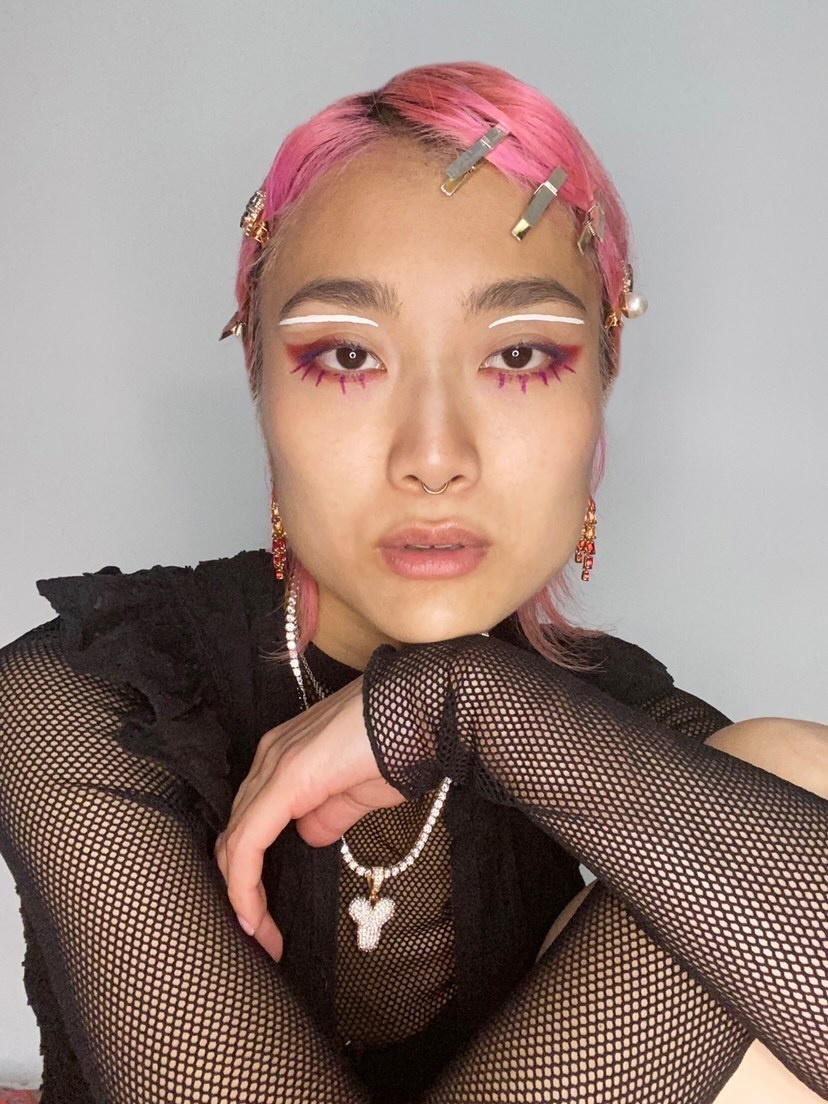 A Japanese person with pink hair slicked down on their head and held in place with clips across their forehead poses with one hand under their chin.