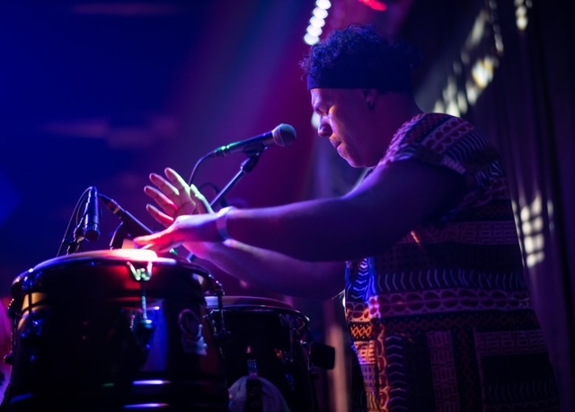 A Puerto Rican man is seen in profile in dramatic purple stage lighting. He is drumming with hands and closes his eyes. 