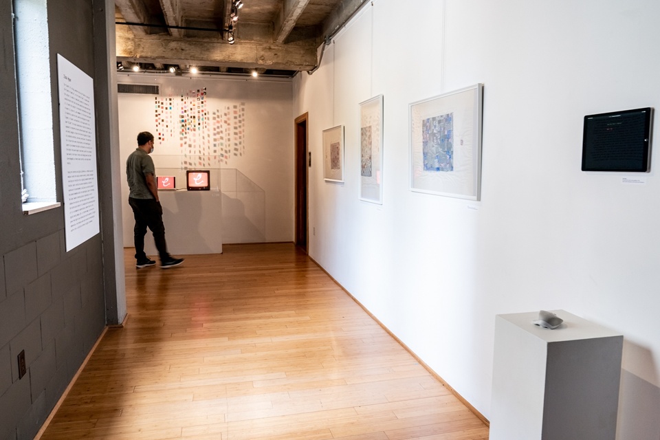 Hallway with three framed artworks and an iPad mounted on the wall. A person looks at a different screen display at the end of the hallway.