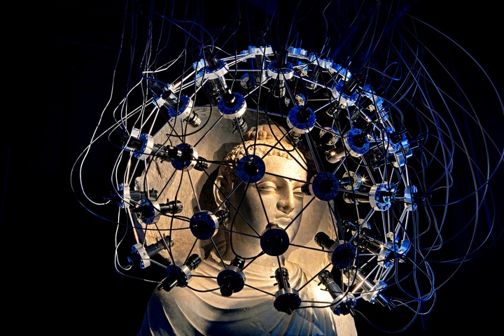 Relief sculptural bust that is yellowish white, surrounded by a circular, interconnect seriees of silver wires and lights giving a slightly bluish hue. The background of the work is black.