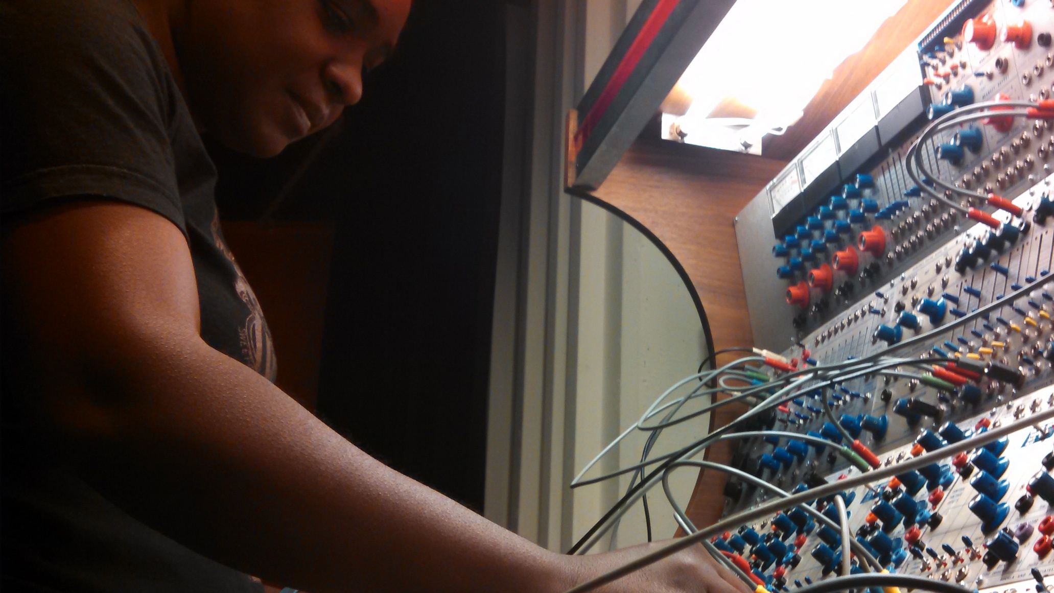 Artist Yvette Janine Jackson stands in front of a sound board with many wires and knobs.
