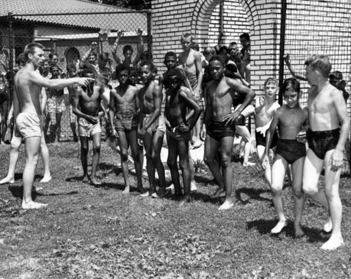A large group of children in swimming suits; one teenager is pointing at them.