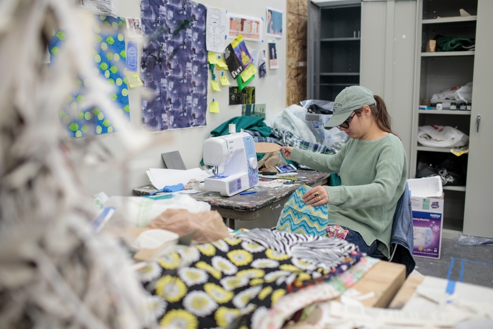 Person works on a fabric project at an L shaped desk covered in fabric scraps and a sewing machine.