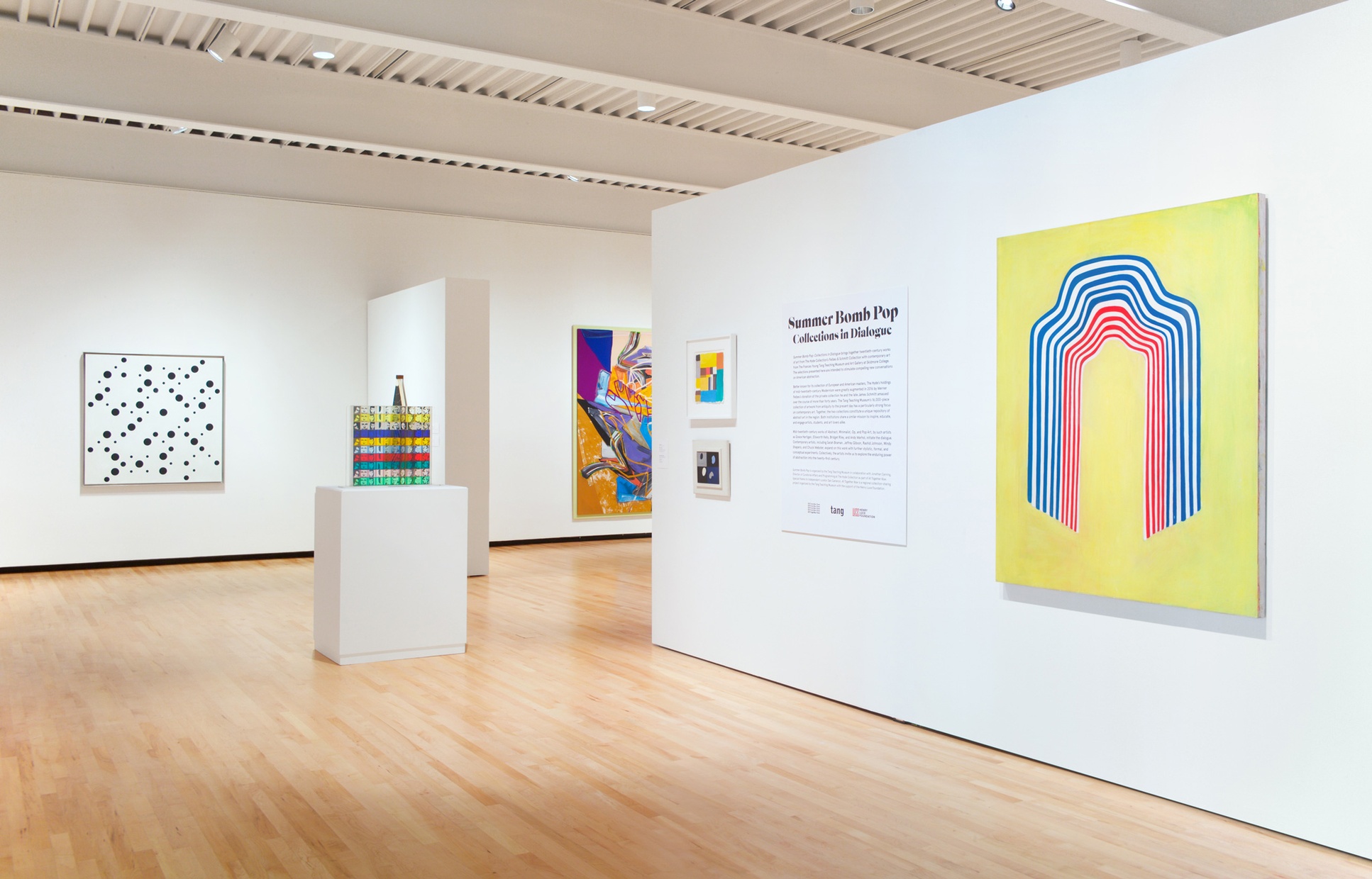 Bright artwork in various mediums is displayed in a gallery with white walls and wood floors. Text on the wall reads "Summer Bomb Pop" with smaller text below.
