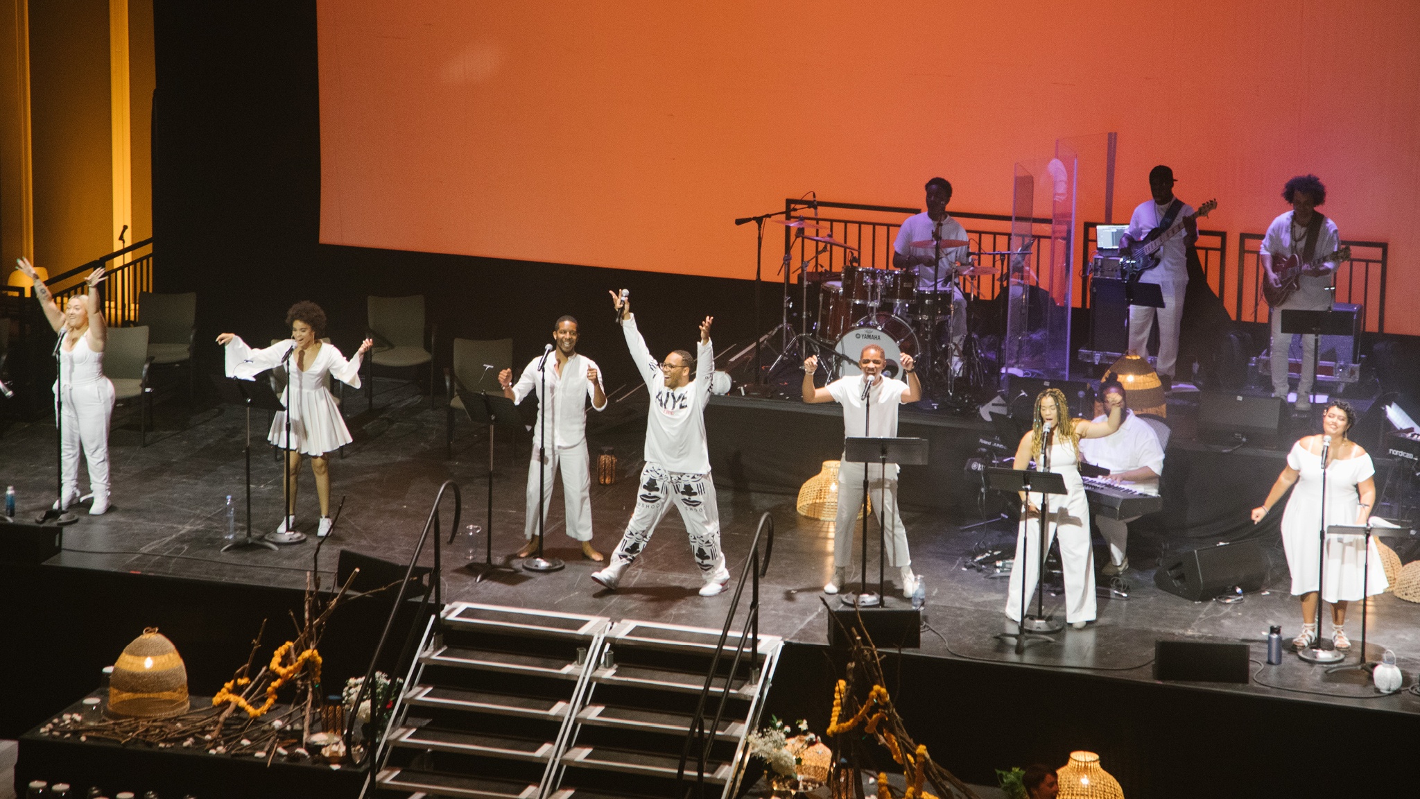 Seven singers performing in a line along the edge of a stage. They all wear white and four backup musicians sit and stand behind them with their instruments.