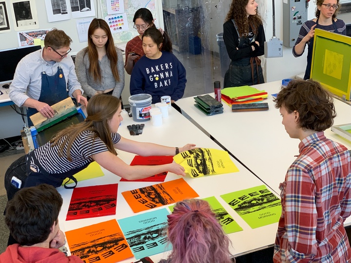 People crowd around a large worktable where colorful silkscreened prayer flags are spread out.