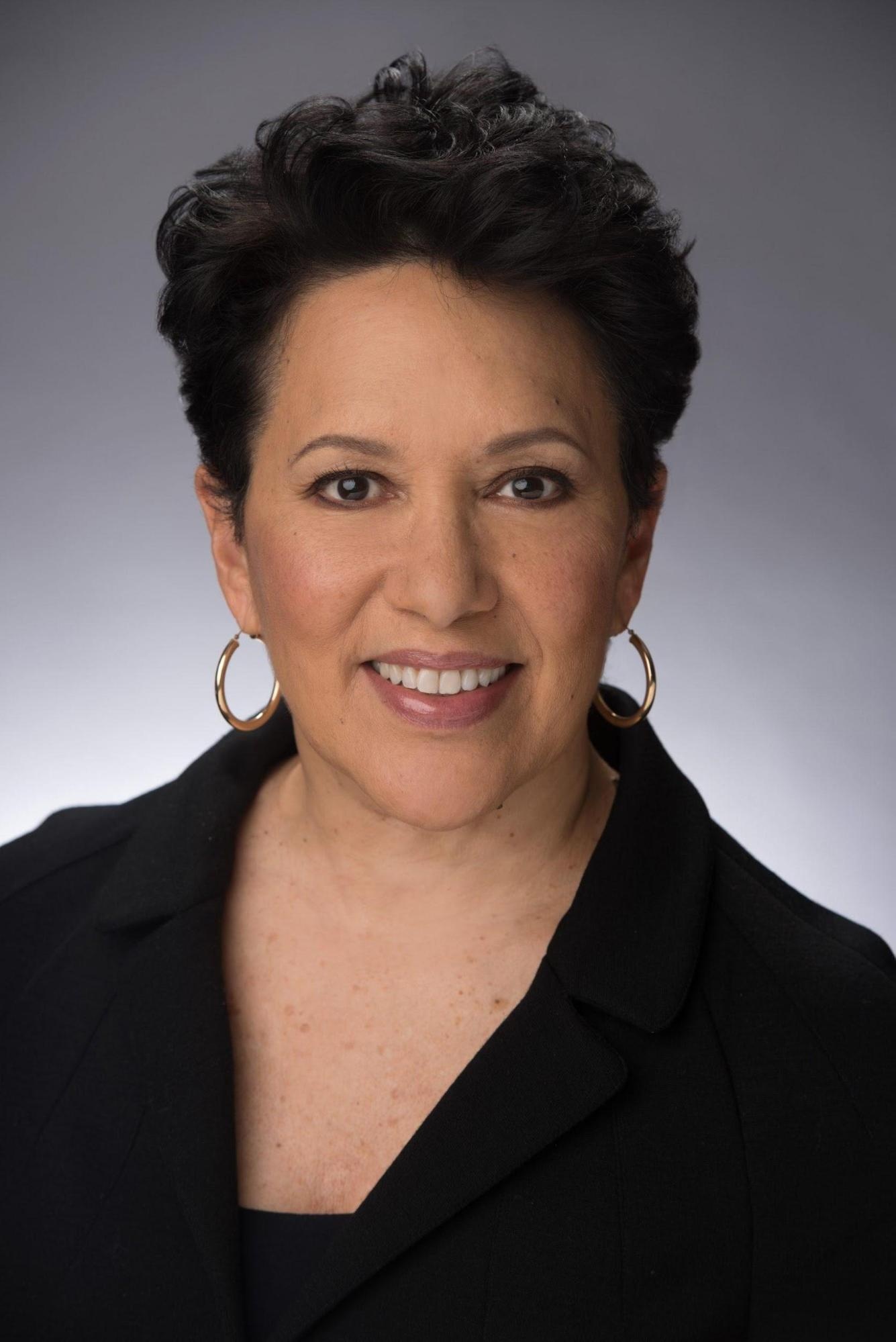 A portrait of cultural worker, producer, and presenter Mikki Shepard, a Black woman with short hair. She wears a black suit jacket and hoop earrings while smiling.