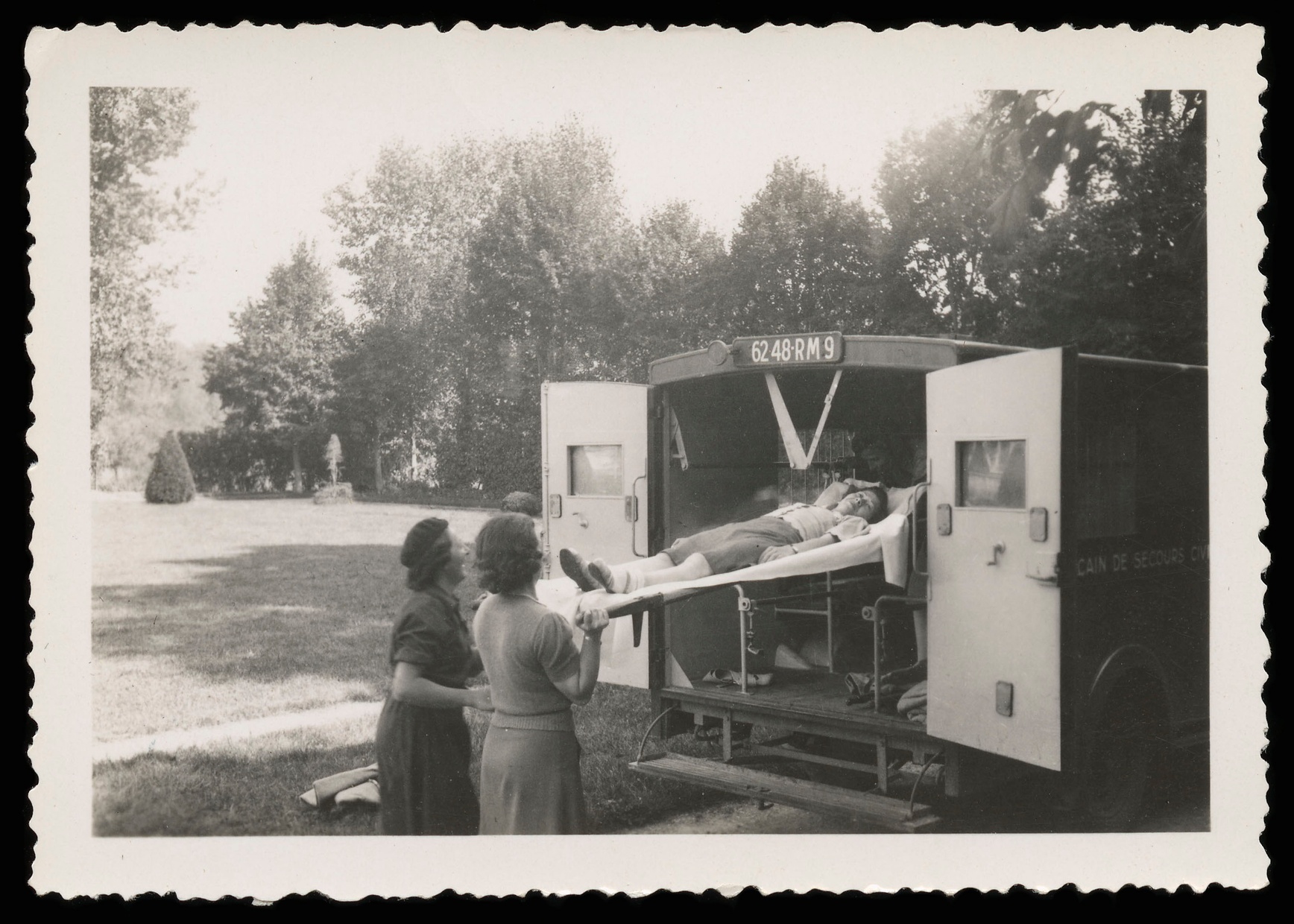 A black and white photograph shows three women at the back of an ambulance. One of them is on a gurney being pushed into the ambulance by the other two women.