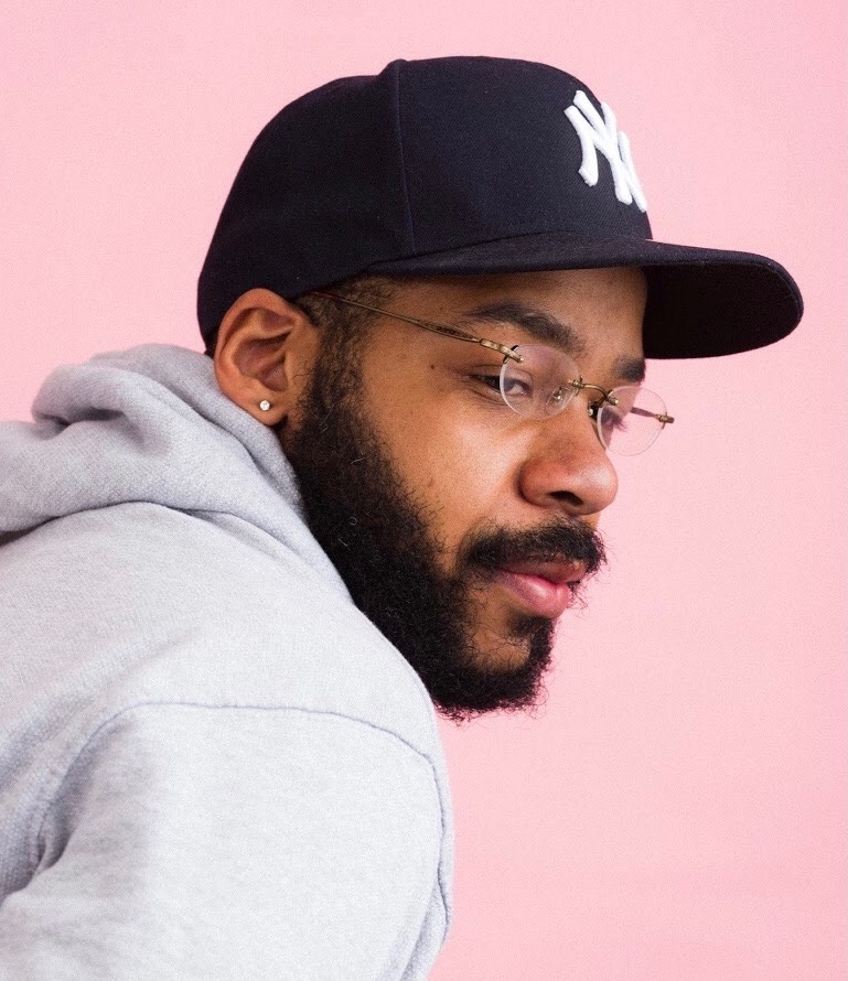 A Black man looks over his shoulder so we see him in three quarters profile. He wears a blue Yankees cap, glasses, and a light gray hoodie. He poses in front of a light pink wall. 