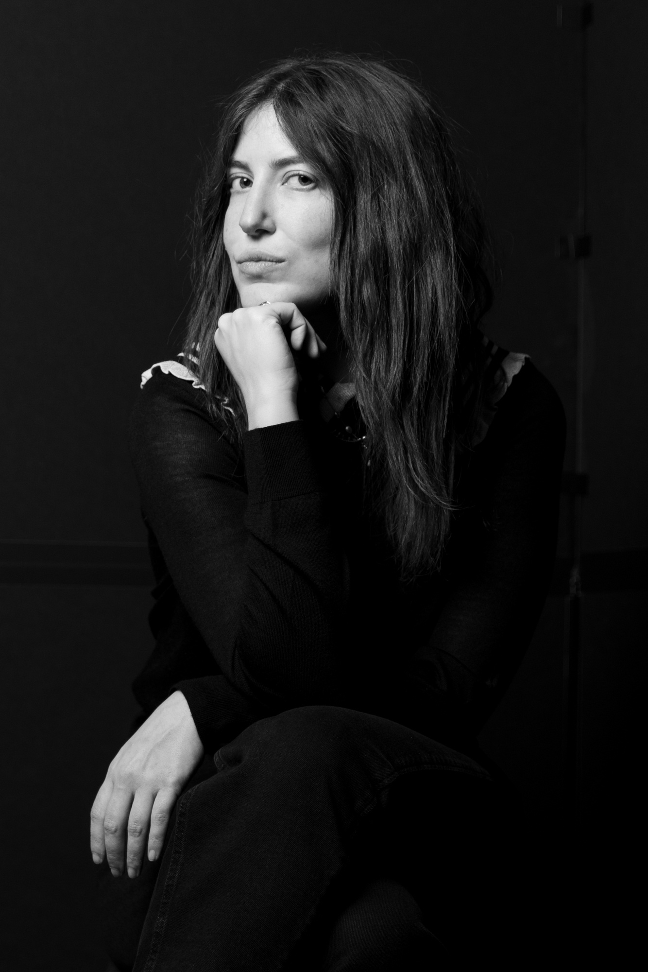 A black and white portrait of Alexandra Rosenberg, who poses with one hand brought to her chin to support her head. She has long dark hair and wears dark clothing that blends in with the dark background. Photo by Maria Baranova