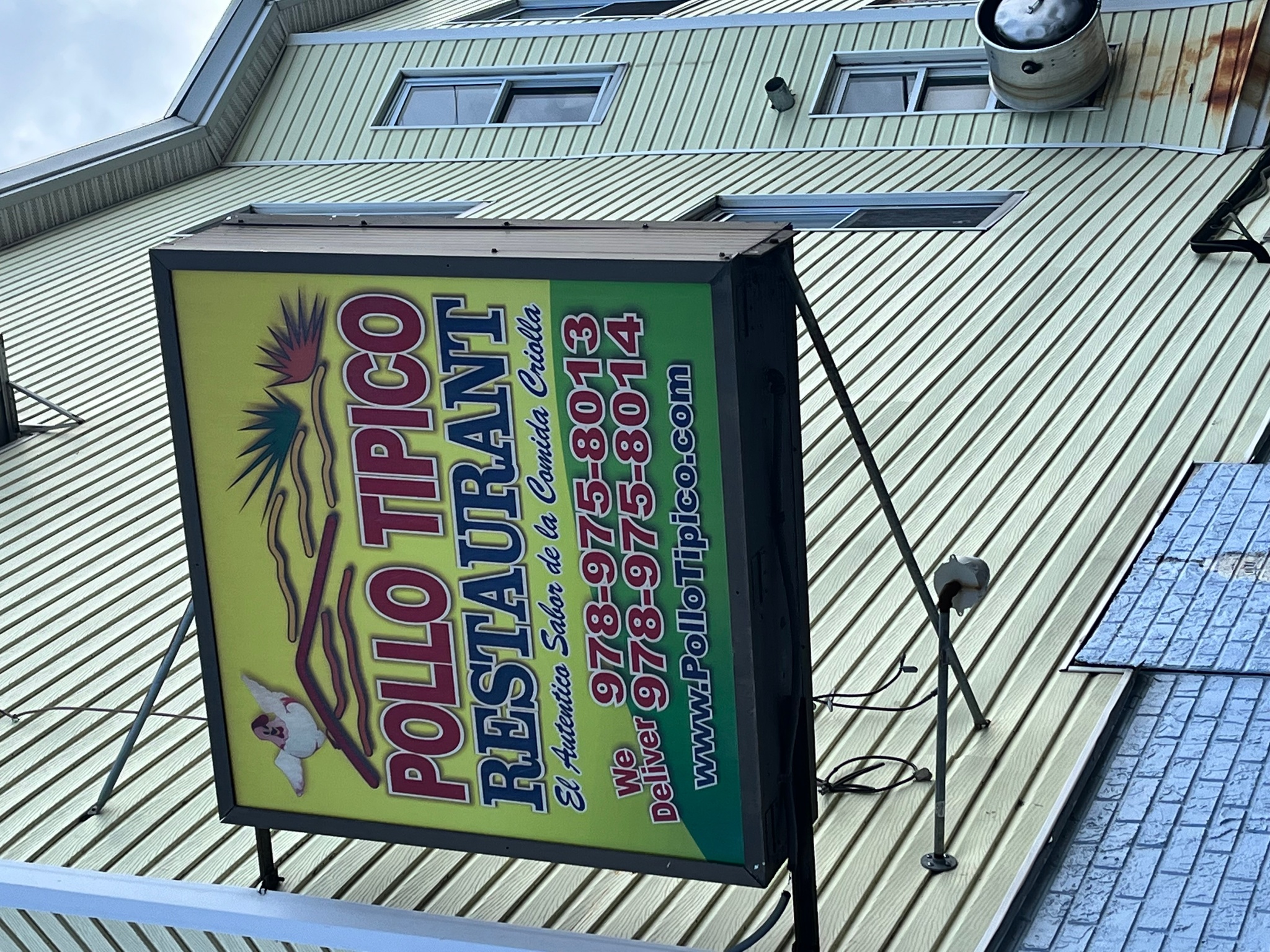 A sign for a Dominican restaurant hanging from the side of a building with yellow siding. The sign reads Pollo Tipico Restaurant.