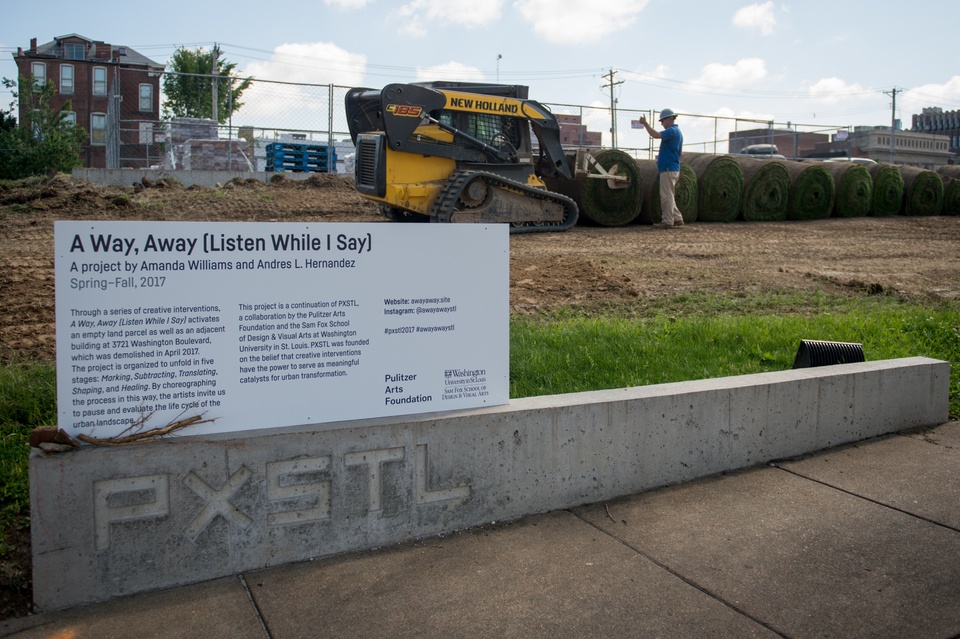 A sign stands in front of a hill with heavy machinery moving around dirt and grass in the background. 