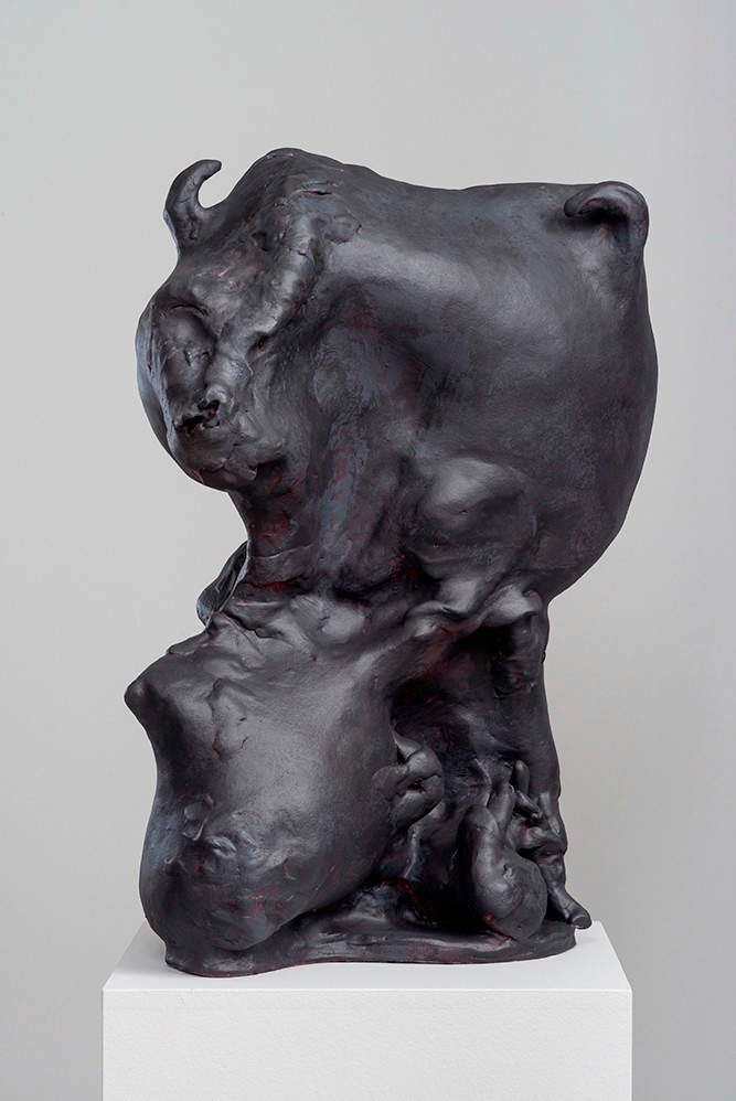 A dark, vaguely amorphous sculpture on a white platform. The sculpture's forms are almost fleshy, with a small horn protruding at the top.