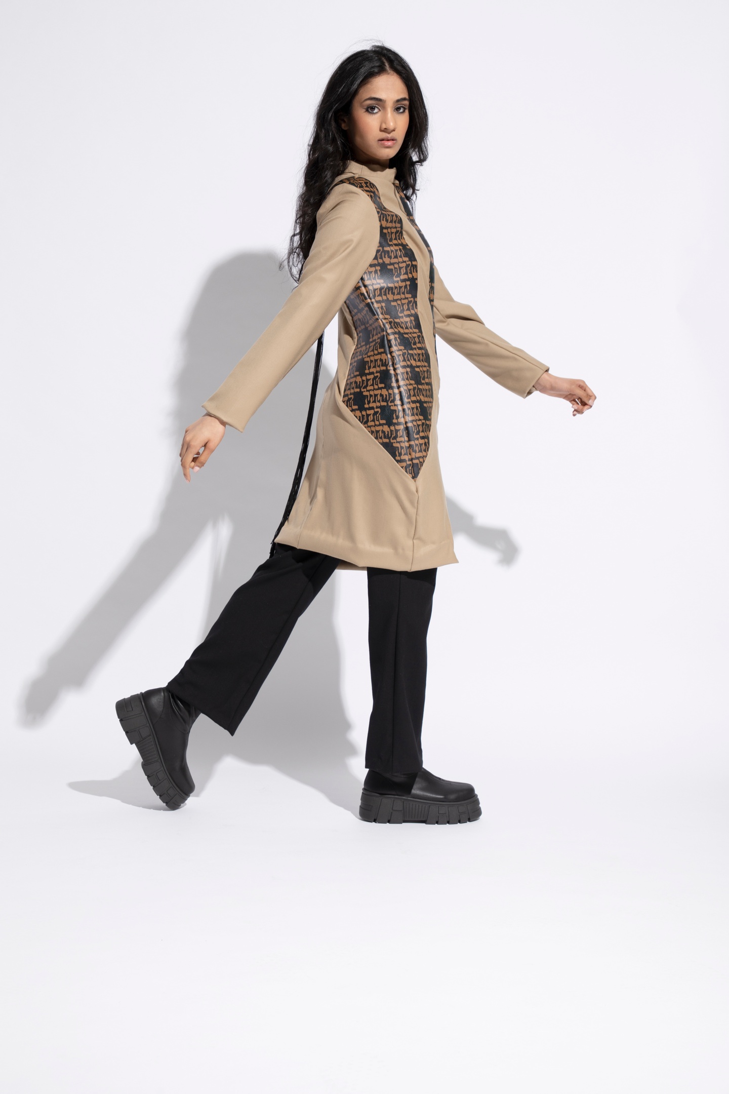 Model wears a beige thigh-length tunic with two side panels running from shoulder to hip made from black leather imprinted with Hebrew characters in sienna. The model wears black bootcut pants and black combat boots.