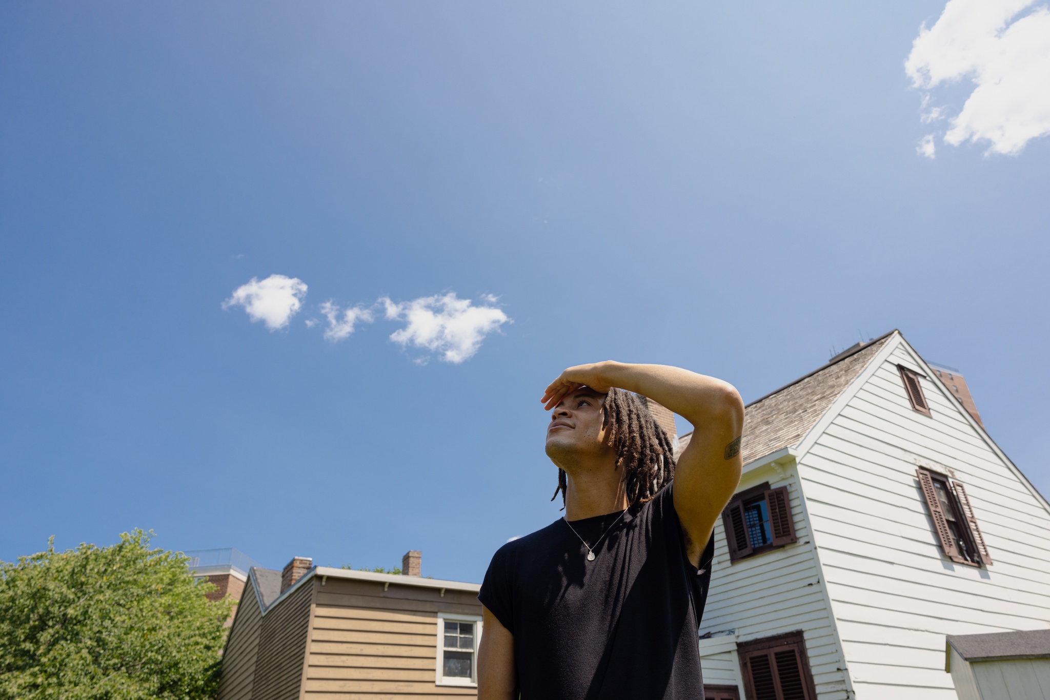 The artist Sandy Williams IV looks up at the blue sky with few clouds above Weeksville Heritage Center. They shield their eyes from the sun.