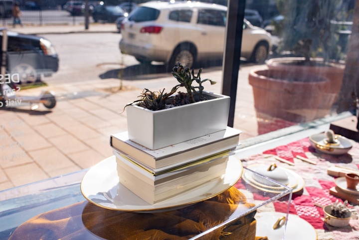 View of an arrangement of dead or dying plants in fancy china displayed on a red and white quilt in a window box looking out onto a city street.