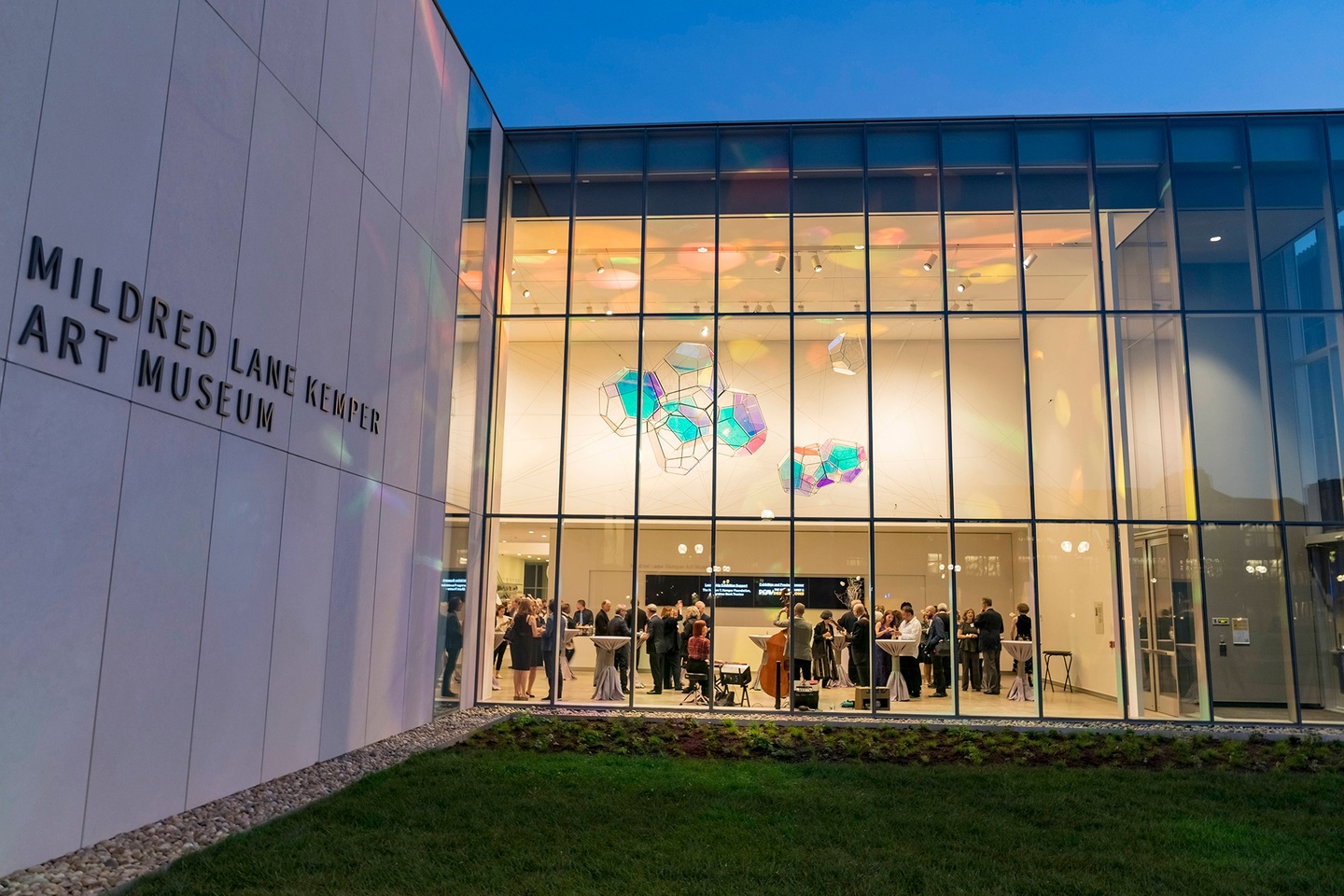 Exterior of the Mildred Lane Kemper Art Museum at night, with lights on and people gathered visible through the large wall of windows