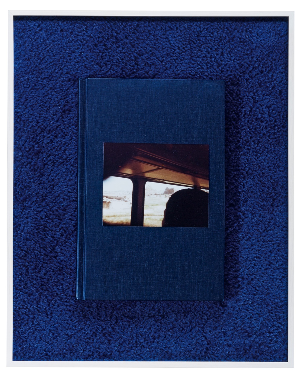 Against a blue carpet sits a blue book with a photograph affixed to the cover depicting a train’s interior with a silhouetted head looking out the window.