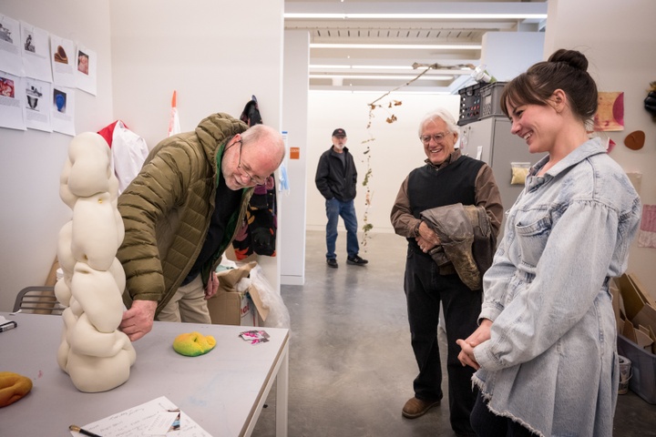 A visitor touches a plaster sculpture on a table in a studio space. The student and another visitor look on, smiling. 