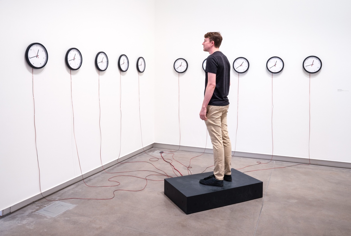 10 clocks on the wall facing a person standing on a black pedestal. There are red wires running from the clocks to the pedestal