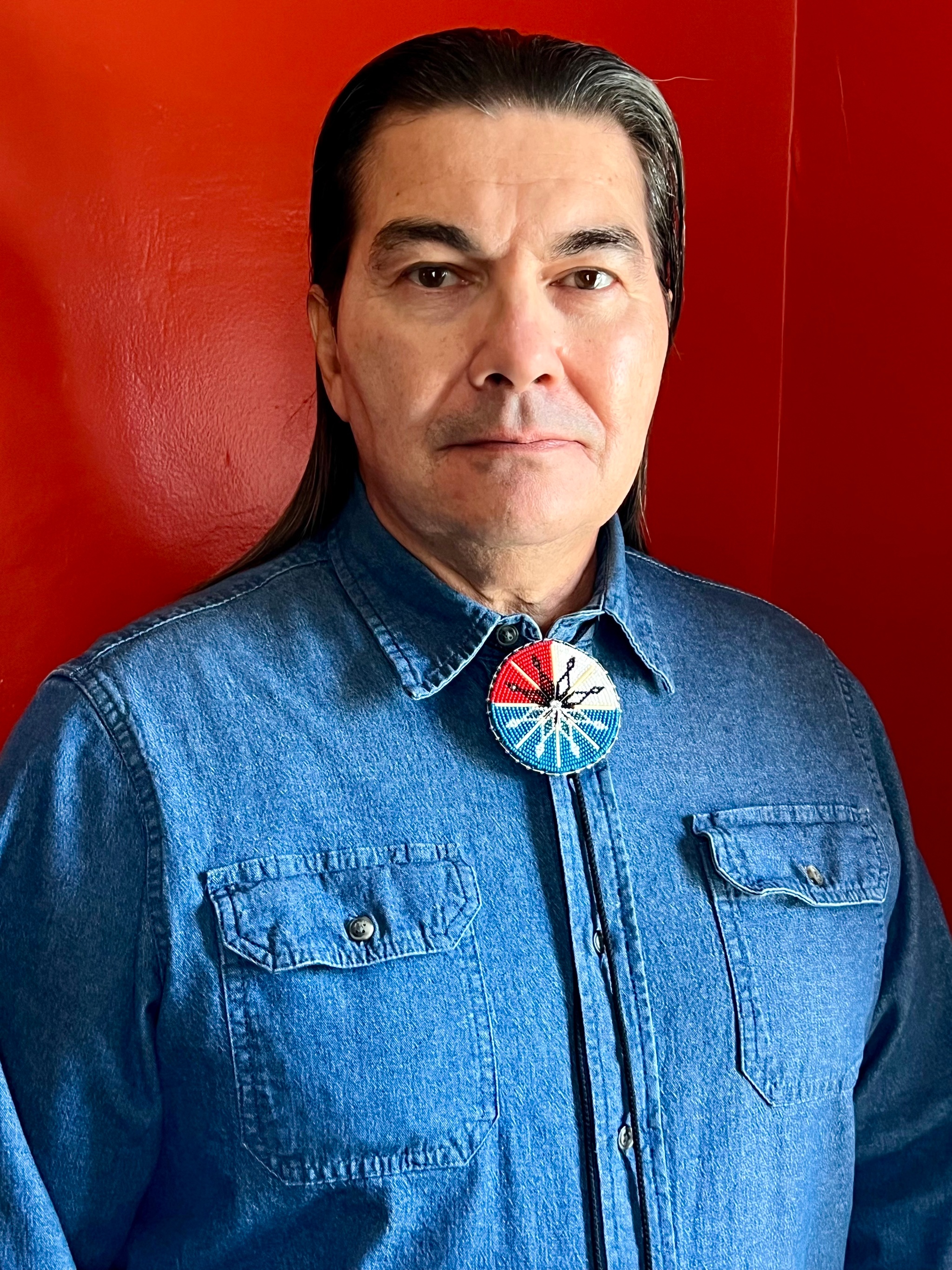 A portrait of Curtis Zunigha, an Indigenous man who poses against a bright red wall. He wears a blue denim collared shirt and has long dark hair that is slicked back behind his head and neck.  