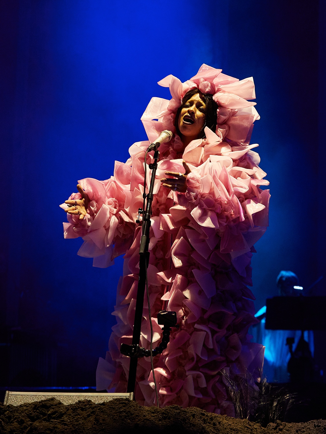 A singer on stage performing in front of a microphone while standing on dirt piled on the stage and wearing a puffy, frilly pink dress with a head covering
