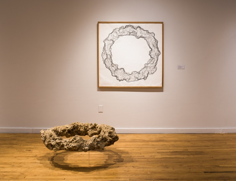 Two works in a gallery; a wall-mounted square wooden frame contains a black and white topographic line drawing of a donut-shaped objects with many lumps and crevices; suspended a few inches off the floor is a sand-colored physical model that matches the drawing.