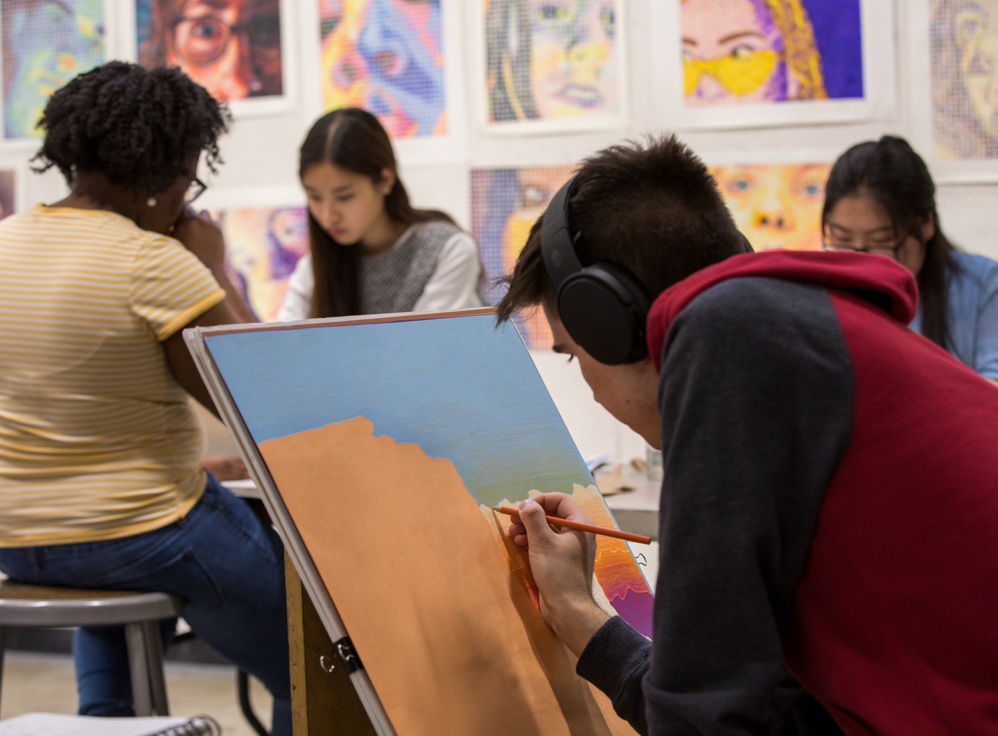 Person in headphones bends over a painting of a desert landscape. Several other people work in the background. The wall is hung with colorful portrait drawings.