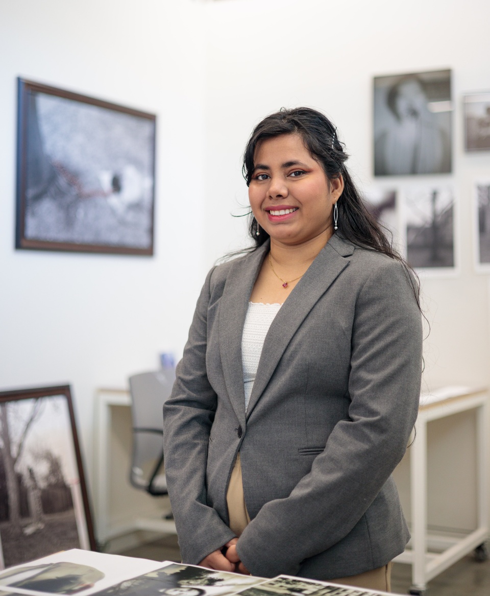 artist Tahia Farhin Haque standing in her studio. She is wearing a gray blazer with white shirt and long hair pulled halfway back. The burred background is the walls covered with in her art