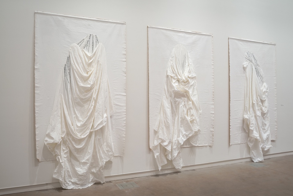 Three large-scale white fabric pieces hung on a white wall. Folds and creases are made in the draped fabric with fine black handstitching.