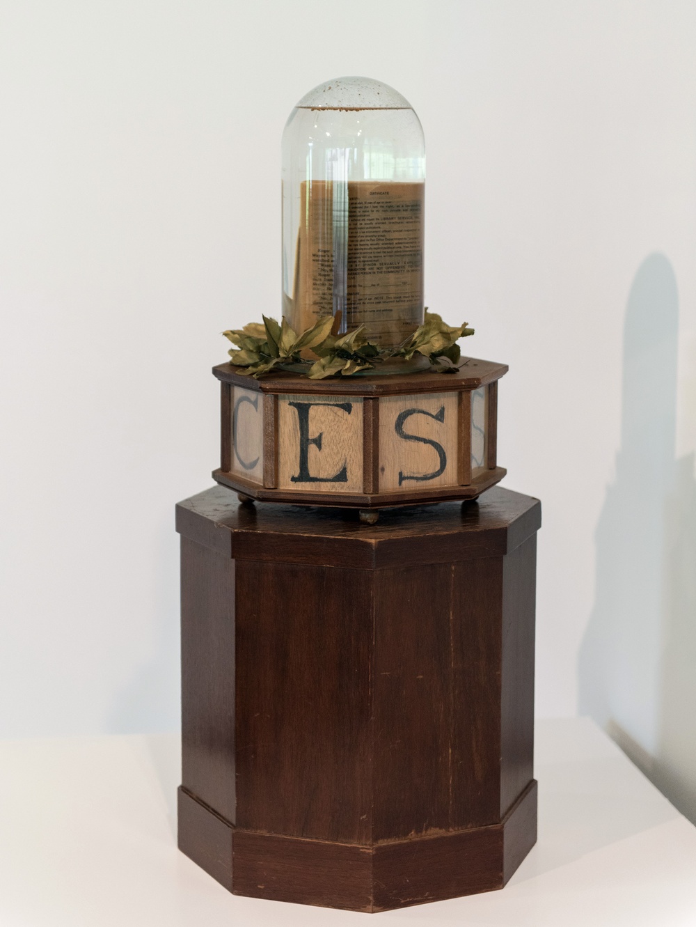 A book submerged in water inside a dome-shaped terrarium is situated on top of a laurel wreath and an octagonal, wooden base with letters  C, E, S