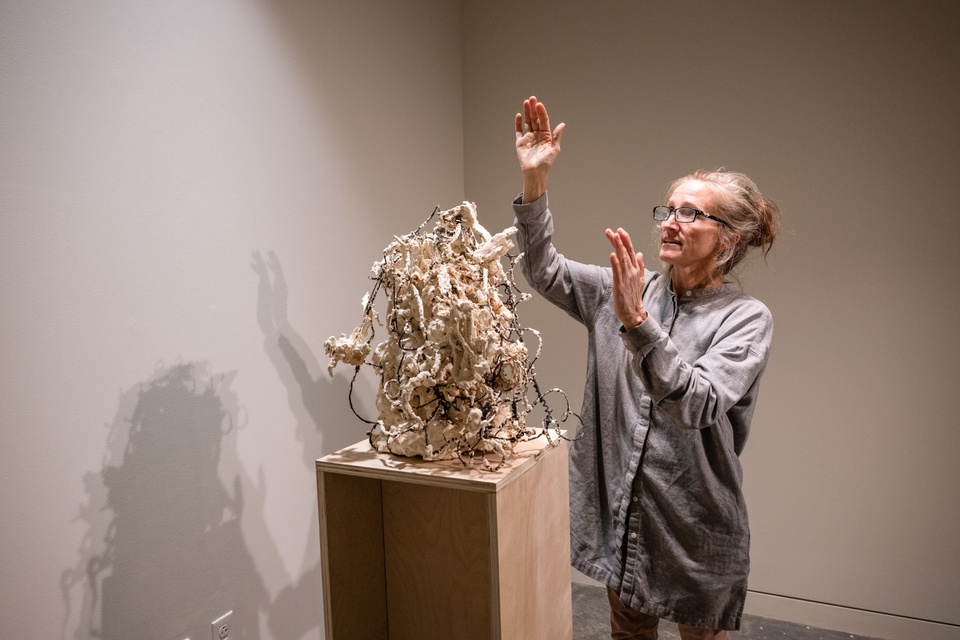 Student stands next to a plinth with a sculpture made of tangled wire coated in plaster blobs and gestures upwards.
