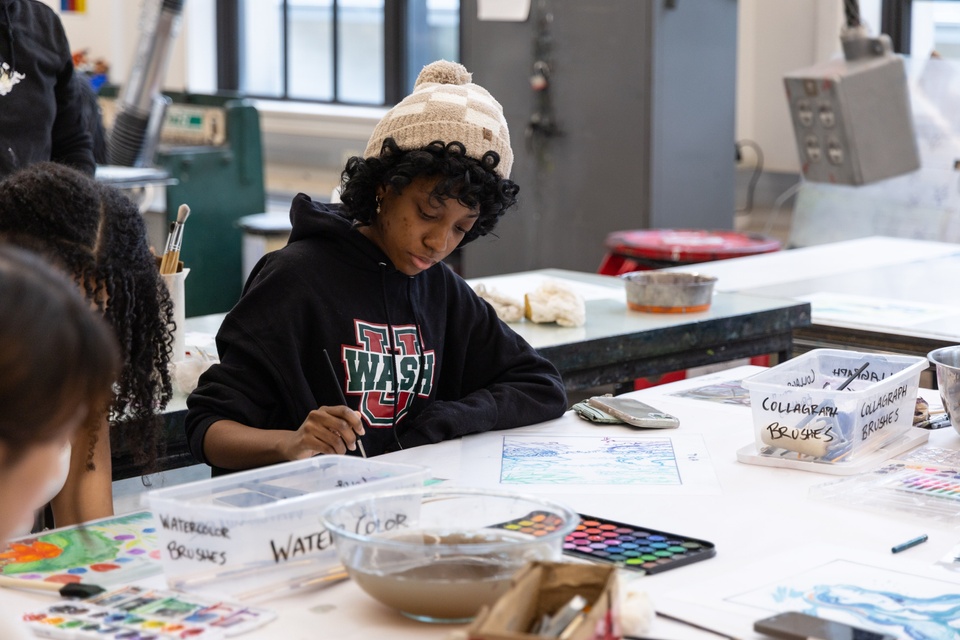 Person seated at a table holds a thin brush and paints on paper. Also on the table are various watercolor palettes and plastic containers containing more brushes.