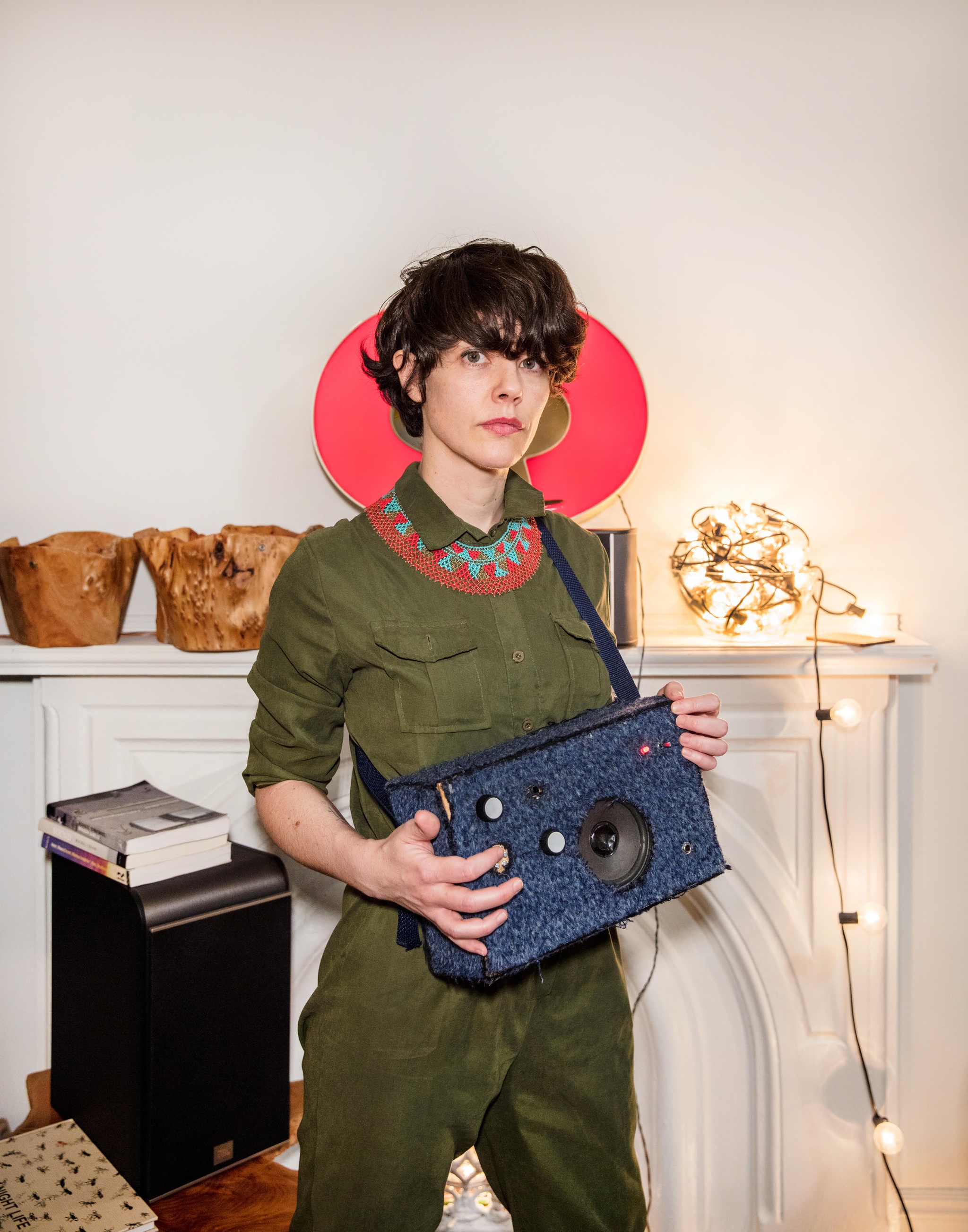 A photo of the artist Merche Blasco who holds a speaker strapped around her torso, with a tangle of string lights in the background on a mantle. 