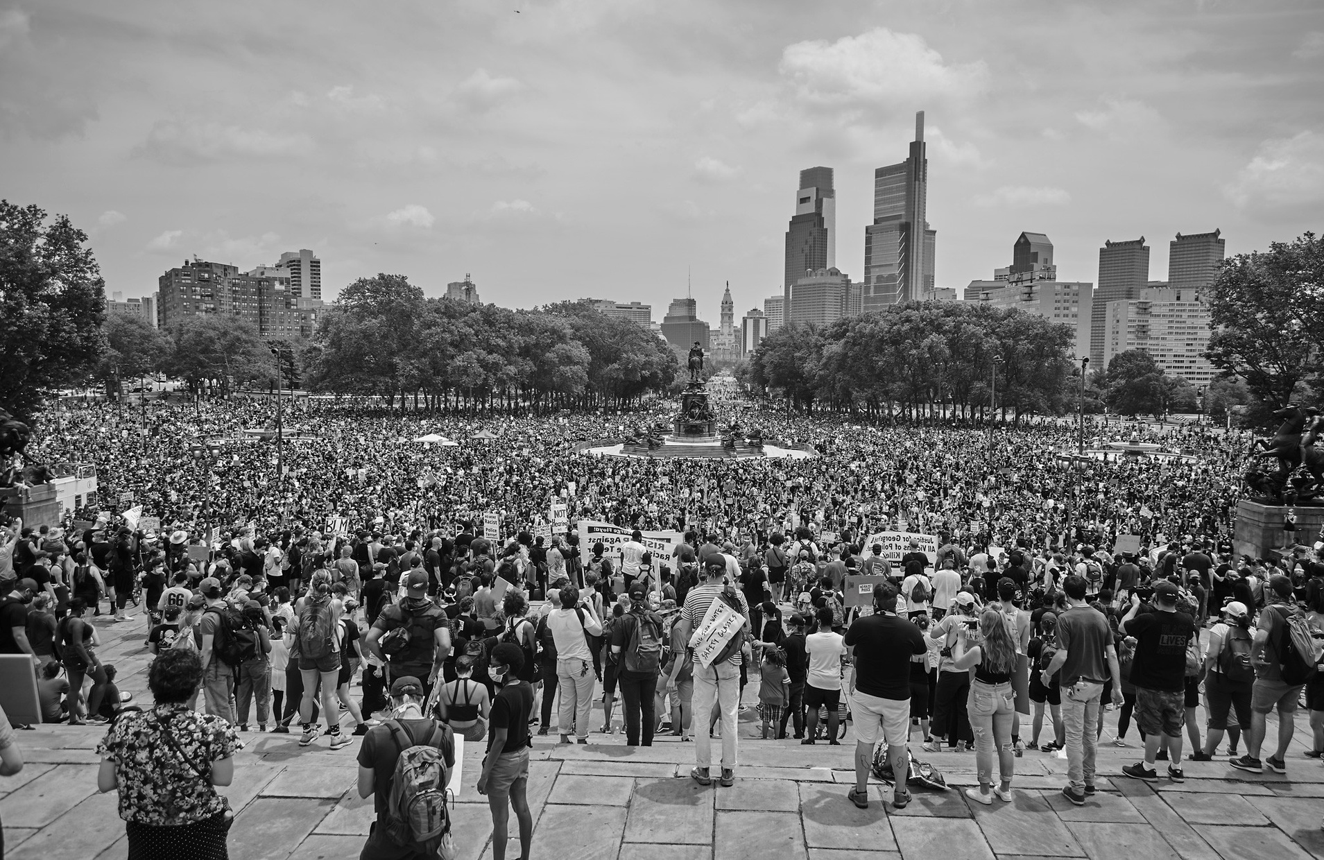 A black and white photograph of a huge crowd of protestors standing facing a cityscape.
