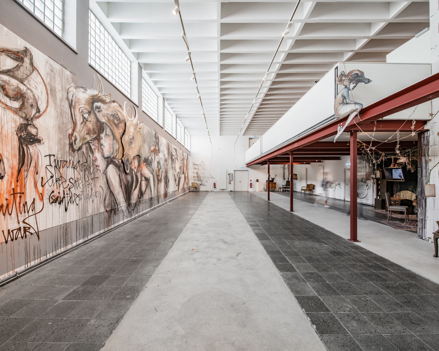 Interior of a museum space with industrial-windows and an iron framework. One wall displays a large-scale painting that is a hybrid of graffiti and cave painting.