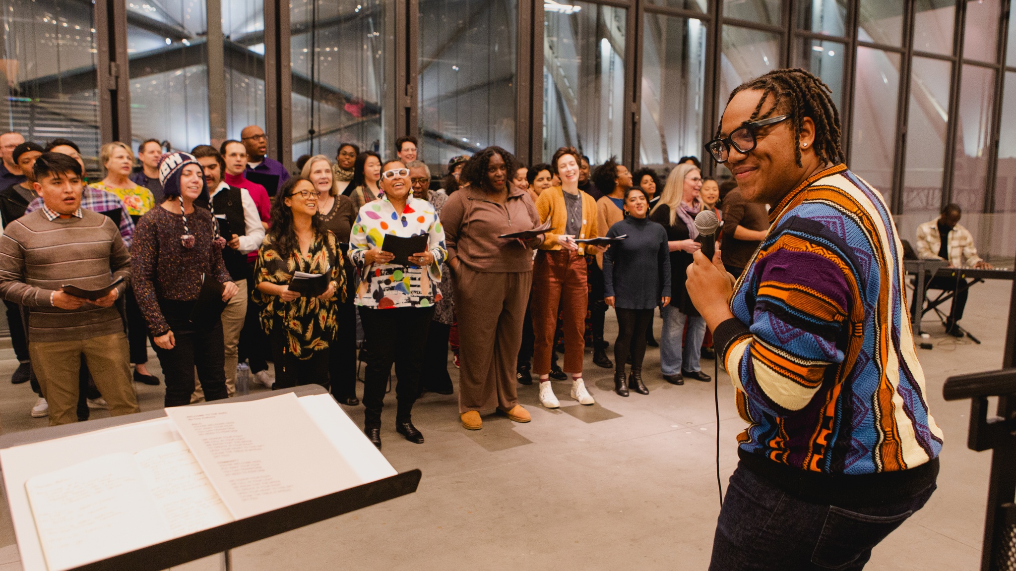 The Fire Ensemble choir rehearses in an event space at The Shed. To the left, the choir's artistic director Troy Anthony stands holding a microphone facing the choir, his head turned to the side with a broad smile on his face. 