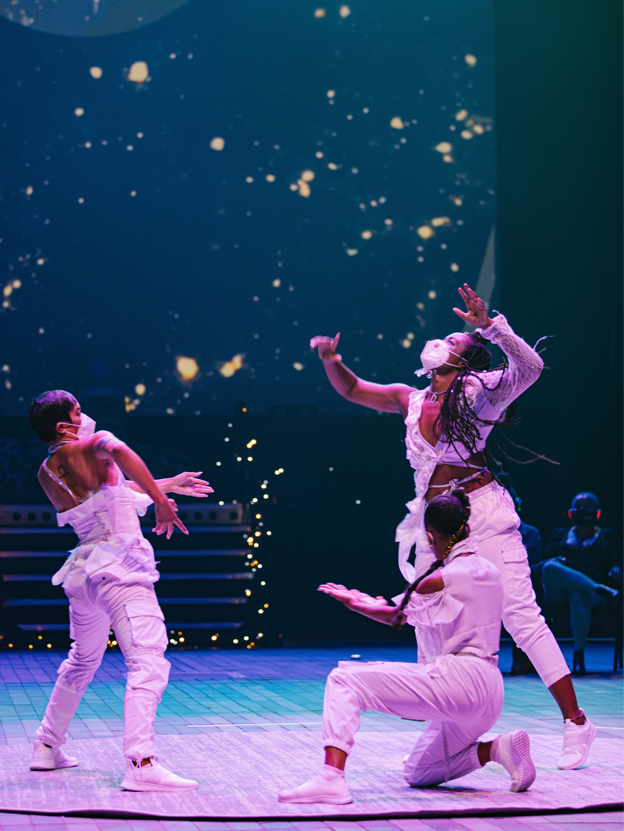 Three performers dressed all in white move on stage with hands out and one of them kneeling. The background scene is lit with small lights evoking a cosmic scene.
