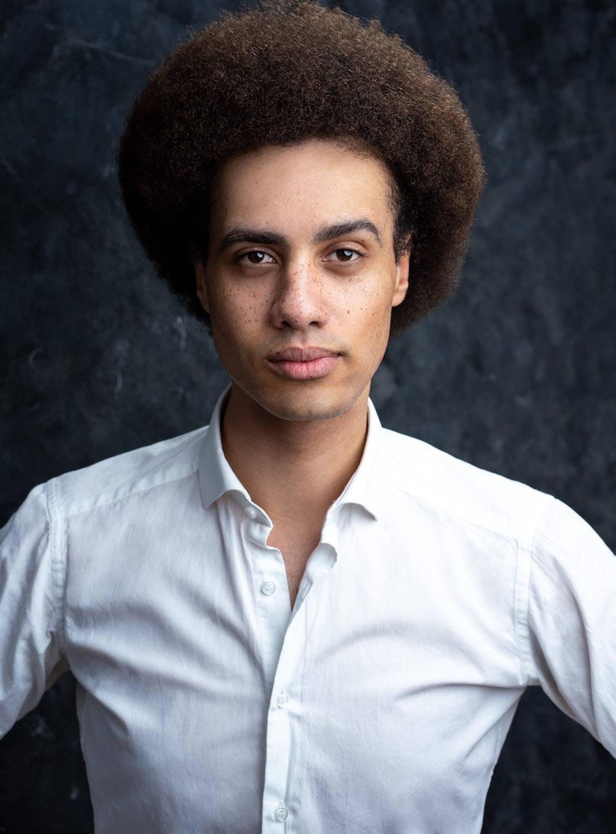 A portrait of Liam Godwin. Liam has an afro and stands with arms out, hands on hips. He looks intently with one eyebrow arched and wears a white button down shirt.