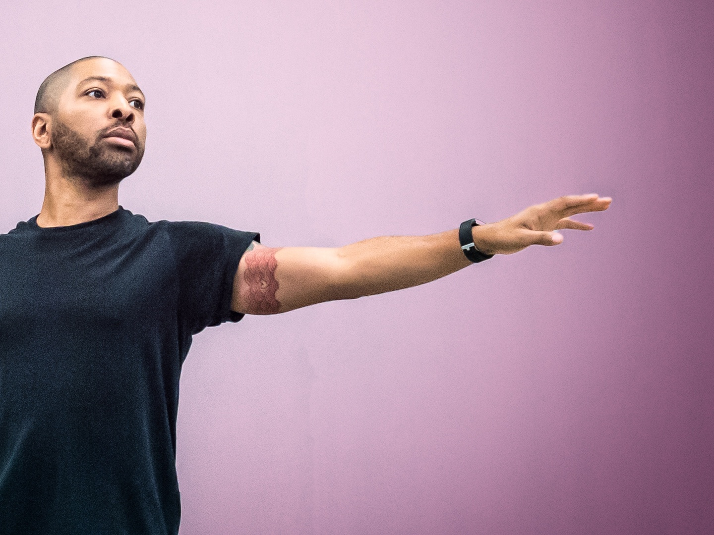 A dark-skinned man stands with his arm extended in front of a pinkish background