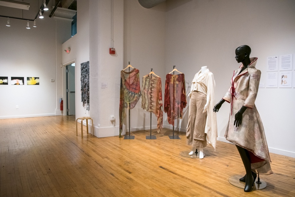 Two mannequins, one wearing a pale coat and the other a canvas tunic with floor-length sleeves, and three dyed robes in autumn colors on hangers are displayed in a gallery space.